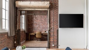 Una habitación con paredes de ladrillo visto, un ventanal con cortinas, suelo de madera, un sofá, un televisor y un escritorio con una silla.