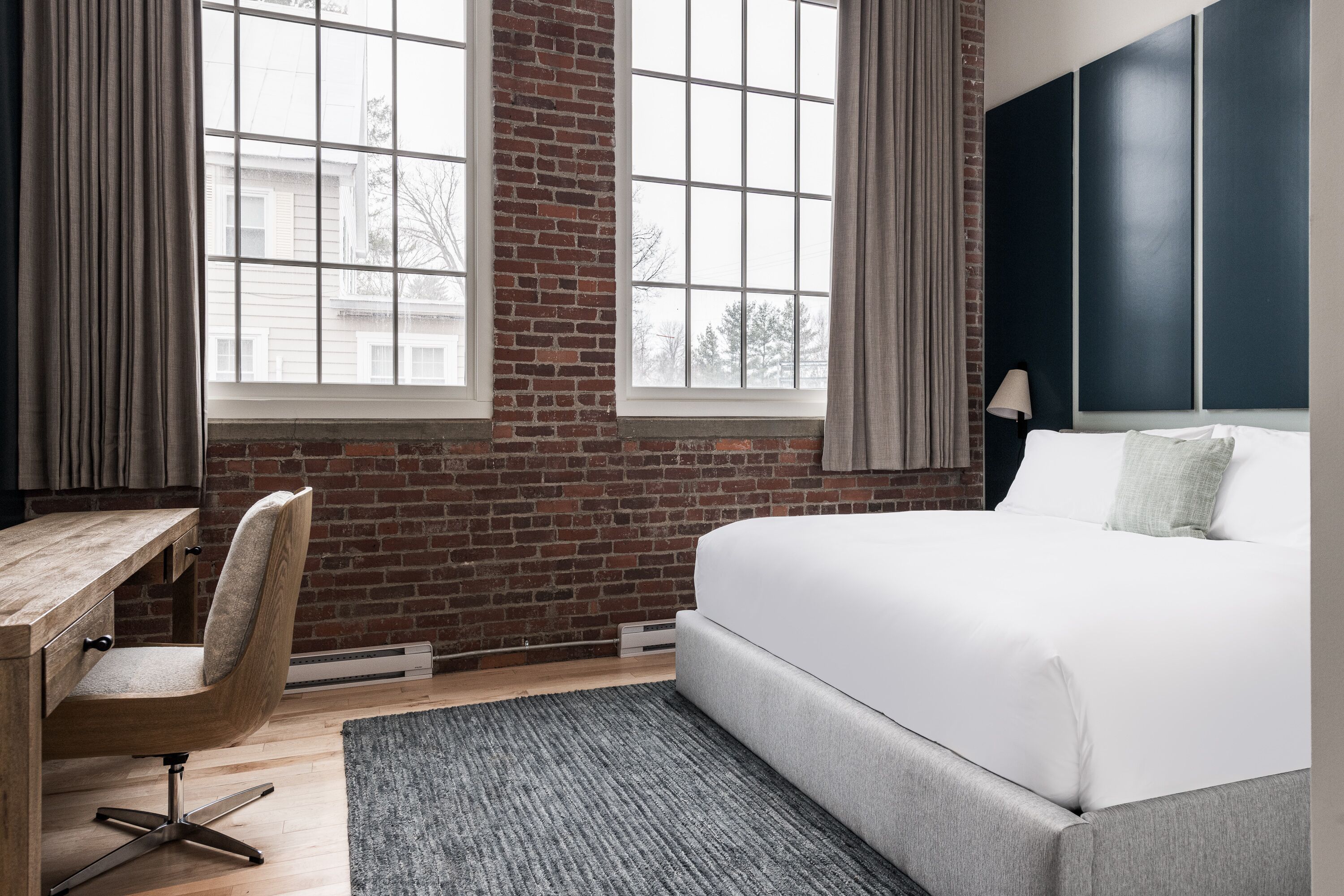 A modern hotel room with a brick wall, large windows, a desk, and a bed.
