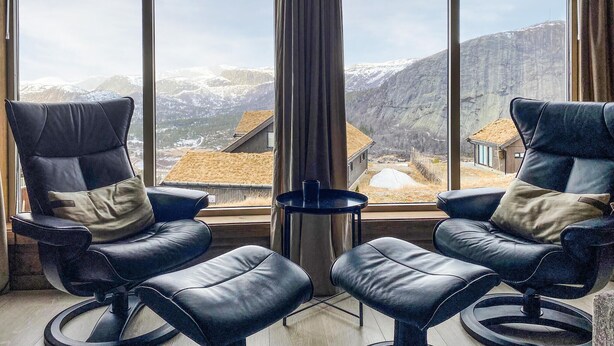 Une pièce avec deux fauteuils en cuir noir et une petite table, de grandes fenêtres offrant une vue sur des montagnes et des maisons enneigées.