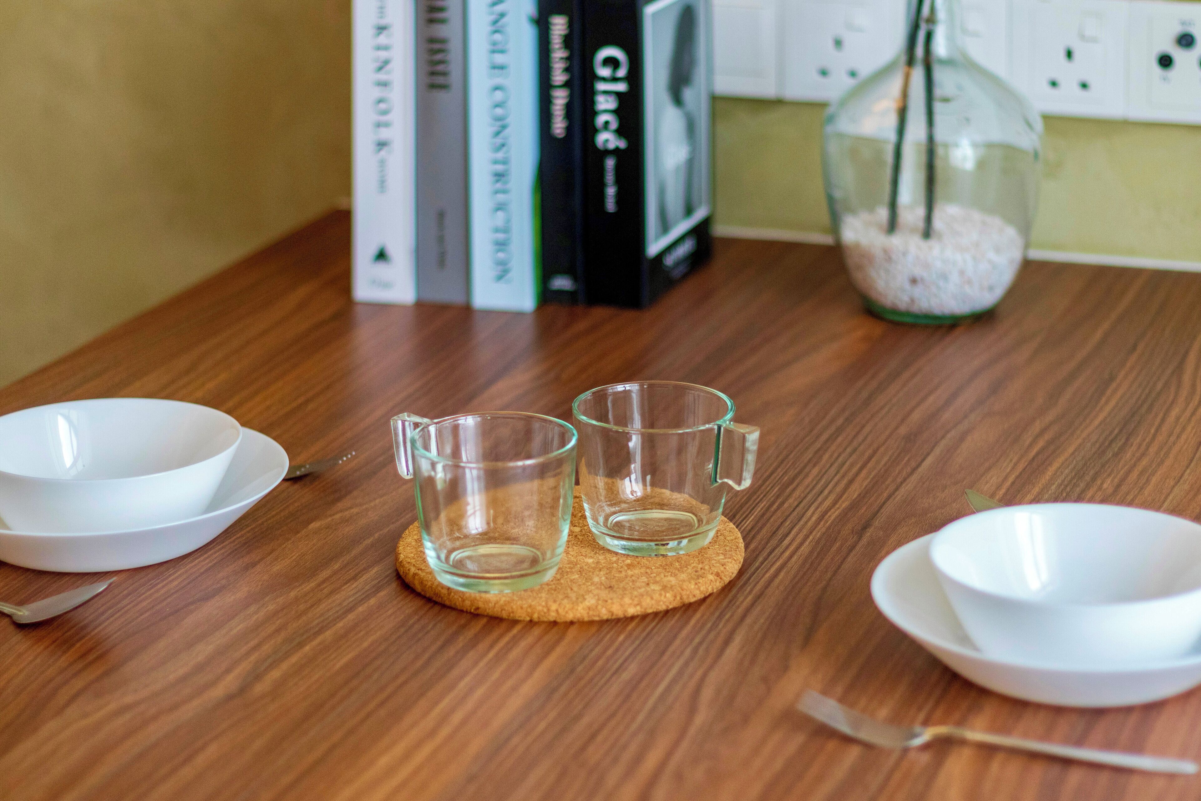 A wooden table with two clear glass cups on a coaster, a white bowl, a spoon, and a glass jar with a plant.