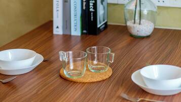 A wooden table with two clear glass cups on a coaster, a white bowl, a spoon, and a glass jar with a plant.