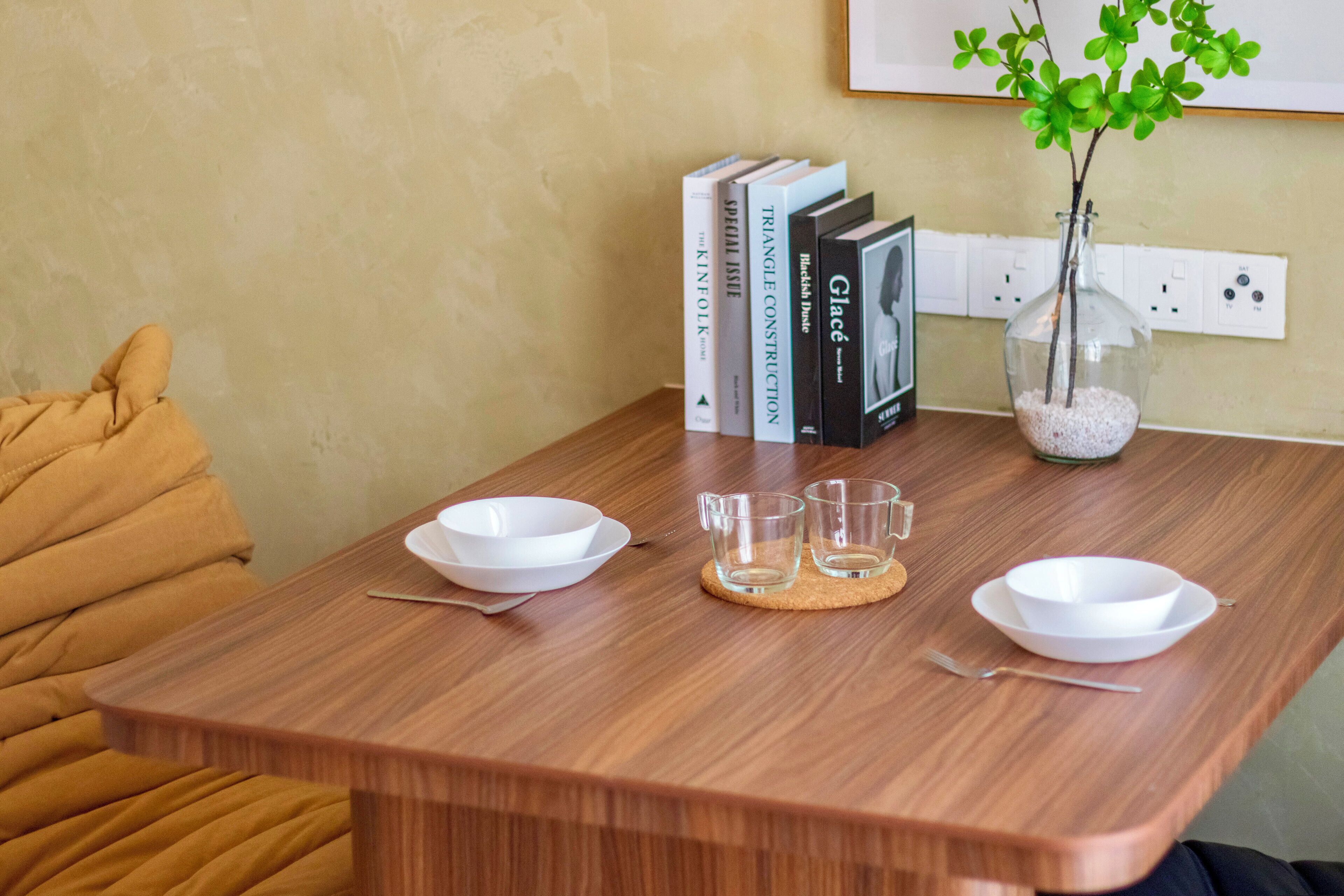 A wooden dining table with two white bowls, two clear glasses, and a small vase with green leaves.