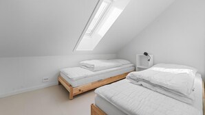 Une petite pièce aux murs blancs et au plafond en pente, comprenant deux lits, une table de chevet et un puits de lumière.