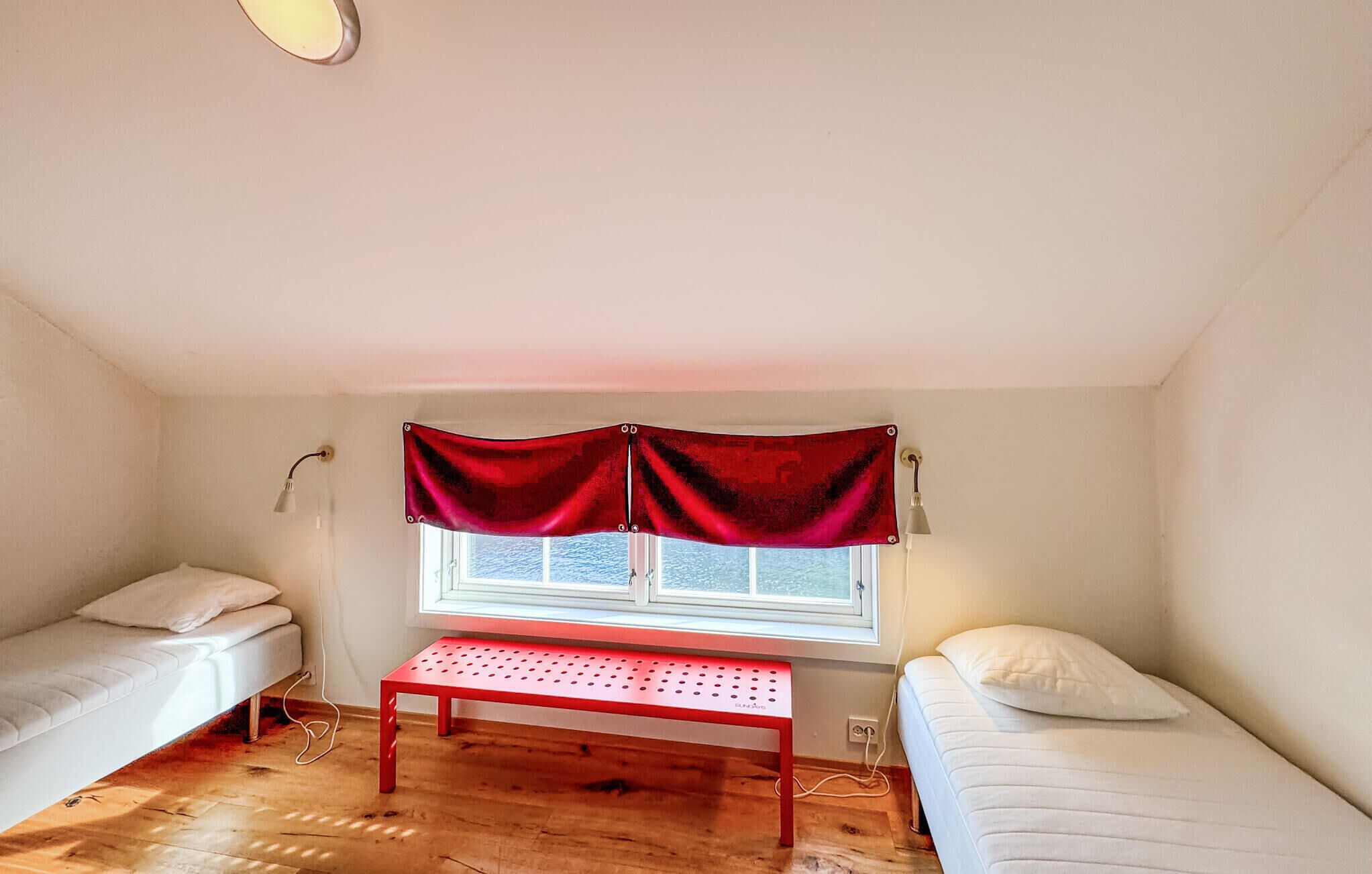 A small, single-room apartment with two single beds, a wooden floor, a window with a red curtain, and a small table.
