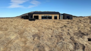 A modern building with large glass windows and a flat roof, surrounded by dry grassland. - Bakkar Luxuary Lodge (Grímsnes- og Grafningshreppur)