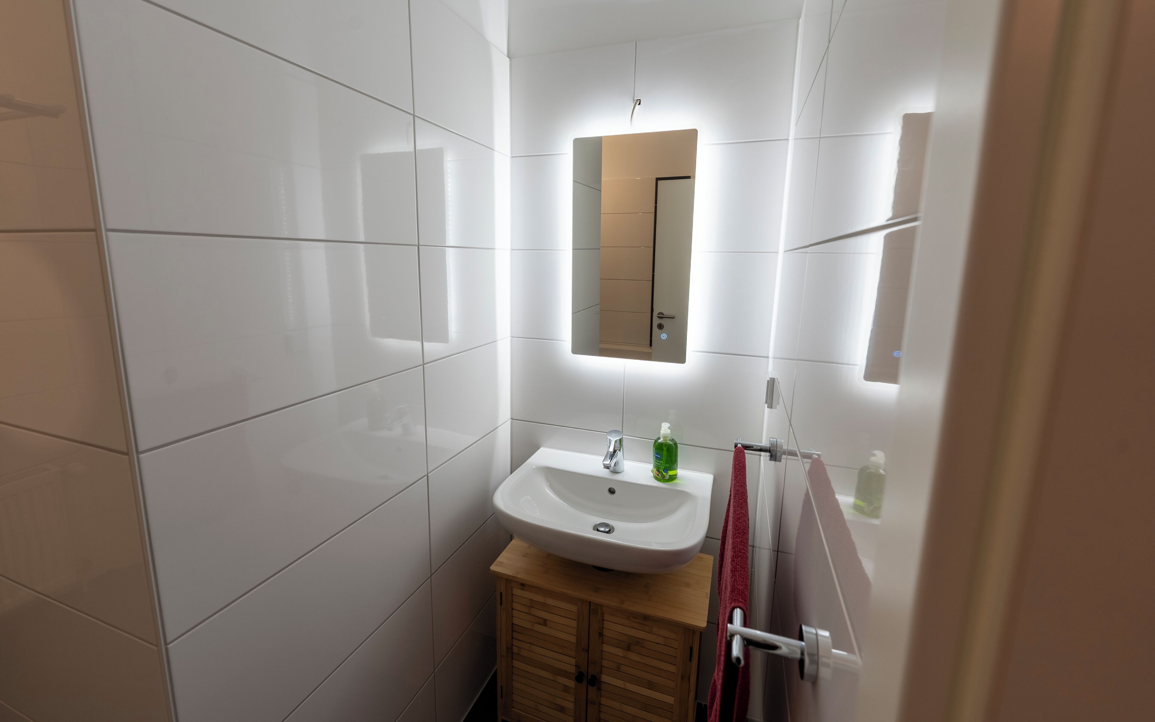 Een moderne badkamer met witte tegels, een wastafel met kraan, een spiegel en een houten kastje.