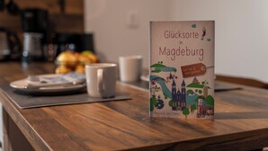 Un livre intitulé « Glücksorte in Magdeburg » est posé sur une table en bois, avec une tasse de café et une assiette de nourriture en arrière-plan.