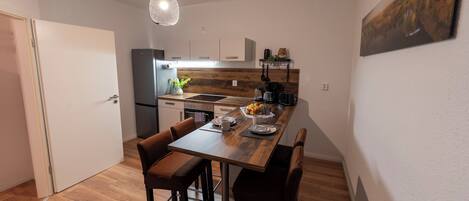 Une cuisine compacte dotée d’un plan de travail en bois, d’un four encastré et d’un réfrigérateur. On y trouve une table à manger avec quatre chaises et un luminaire suspendu.