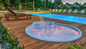 A hot tub on a wooden deck with lounge chairs and a pool in the background.