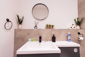 A modern bathroom with a white sink, black faucet, and a round mirror on the wall. There are potted plants and toiletries on the vanity. - City Getaway: Cosy 1Bed Apt in Hull Centre (Hull)
