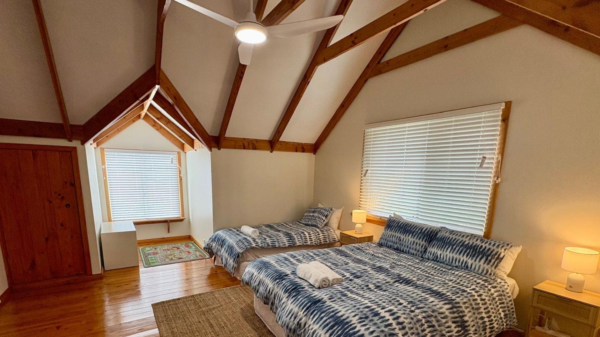 Une chambre avec deux lits, des poutres en bois, un ventilateur de plafond et une fenêtre avec des stores.