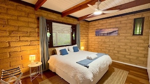 A bedroom with a large bed, a ceiling fan, a window with blinds, and a brick wall. - Timber Tides Retreat (Geographe)