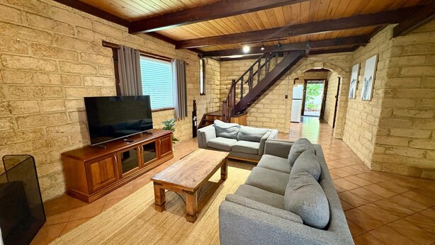 A spacious living room with a wooden TV stand, a grey sofa, and a wooden coffee table. A staircase leads to an upper level. - Timber Tides Retreat (Geographe)