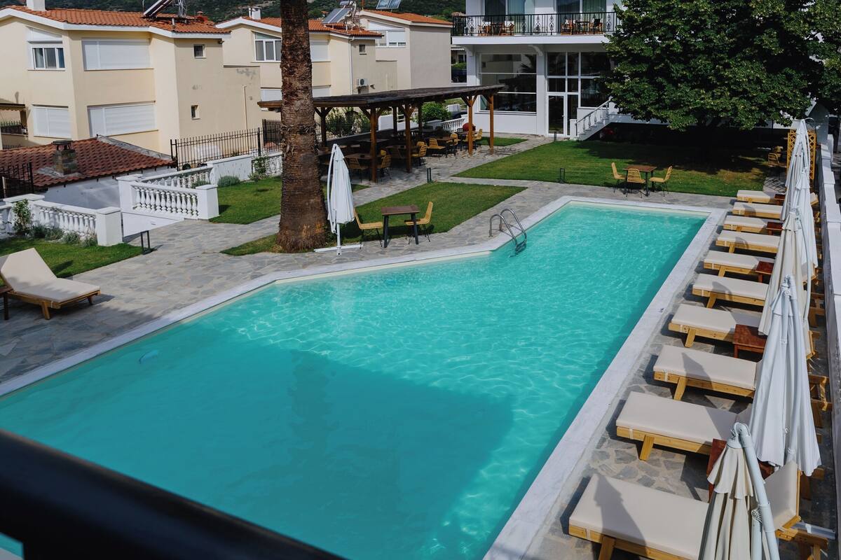 Ein Swimmingpool mit Liegestühlen und Sonnenschirmen, umgeben von einem angelegten Garten und Gebäuden.