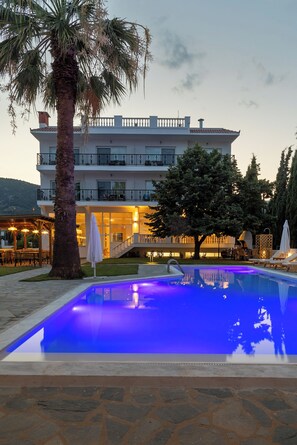 Una piscina iluminada con luces azules, una palmera y un edificio con balcones y terraza.