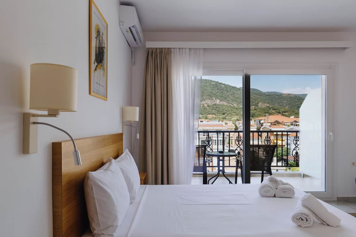 Ein Hotelzimmer mit hölzernem Kopfteil, einem Bett mit weißer Bettwäsche, einer Nachttischlampe und Blick auf einen Balkon mit Tisch und Stühlen. Durch das Fenster ist eine Berglandschaft sichtbar.