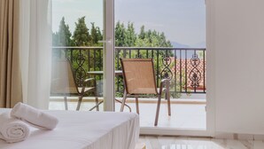 A hotel room with a bed, a balcony with a chair, and a view of greenery and mountains.