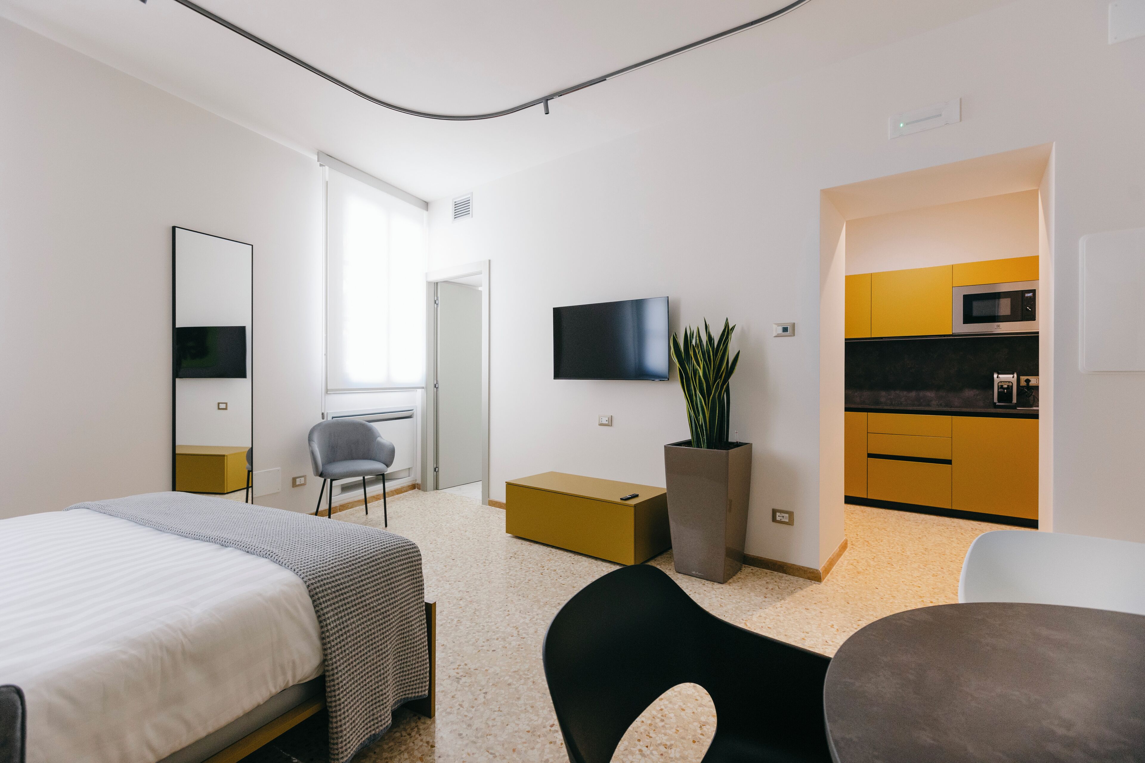 A modern hotel room with a bed, a desk, a chair, a TV, a plant, and a kitchenette.