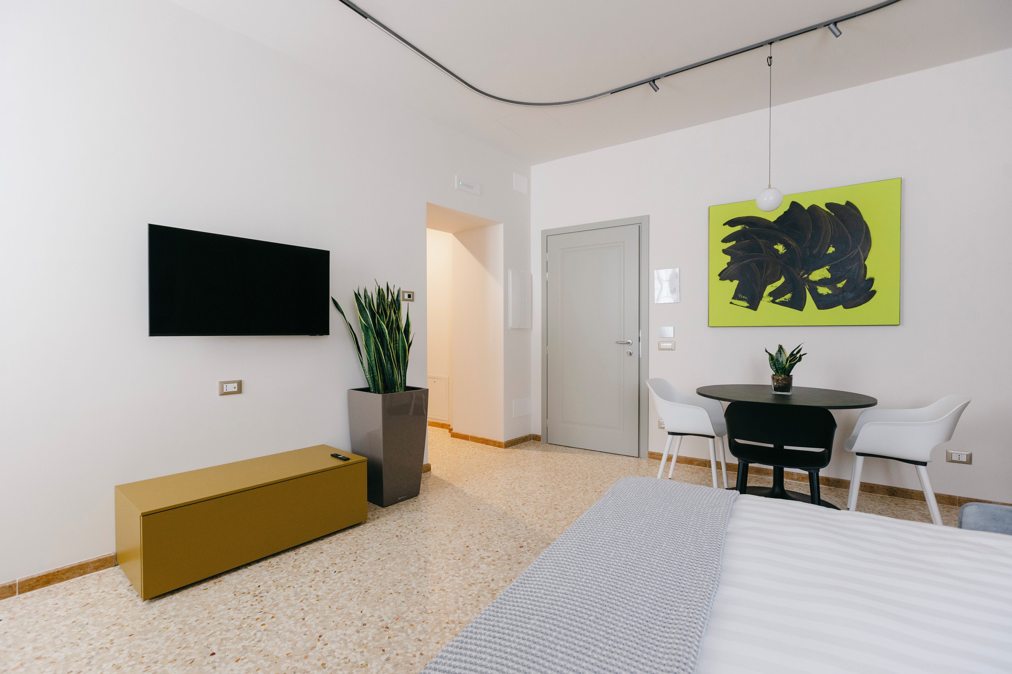 A modern hotel room with a bed, a desk with a chair, a flat-screen TV mounted on the wall, a potted plant, and a painting.