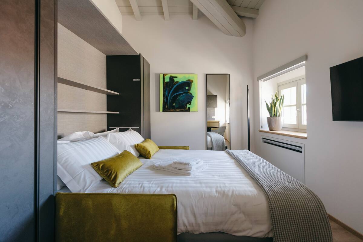 Ein modernes Hotelzimmer mit einem ordentlich bezogenen Bett, einem mit Stoff gepolsterten grünen Sessel und einem großen Fenster mit einem Blumentopf.
