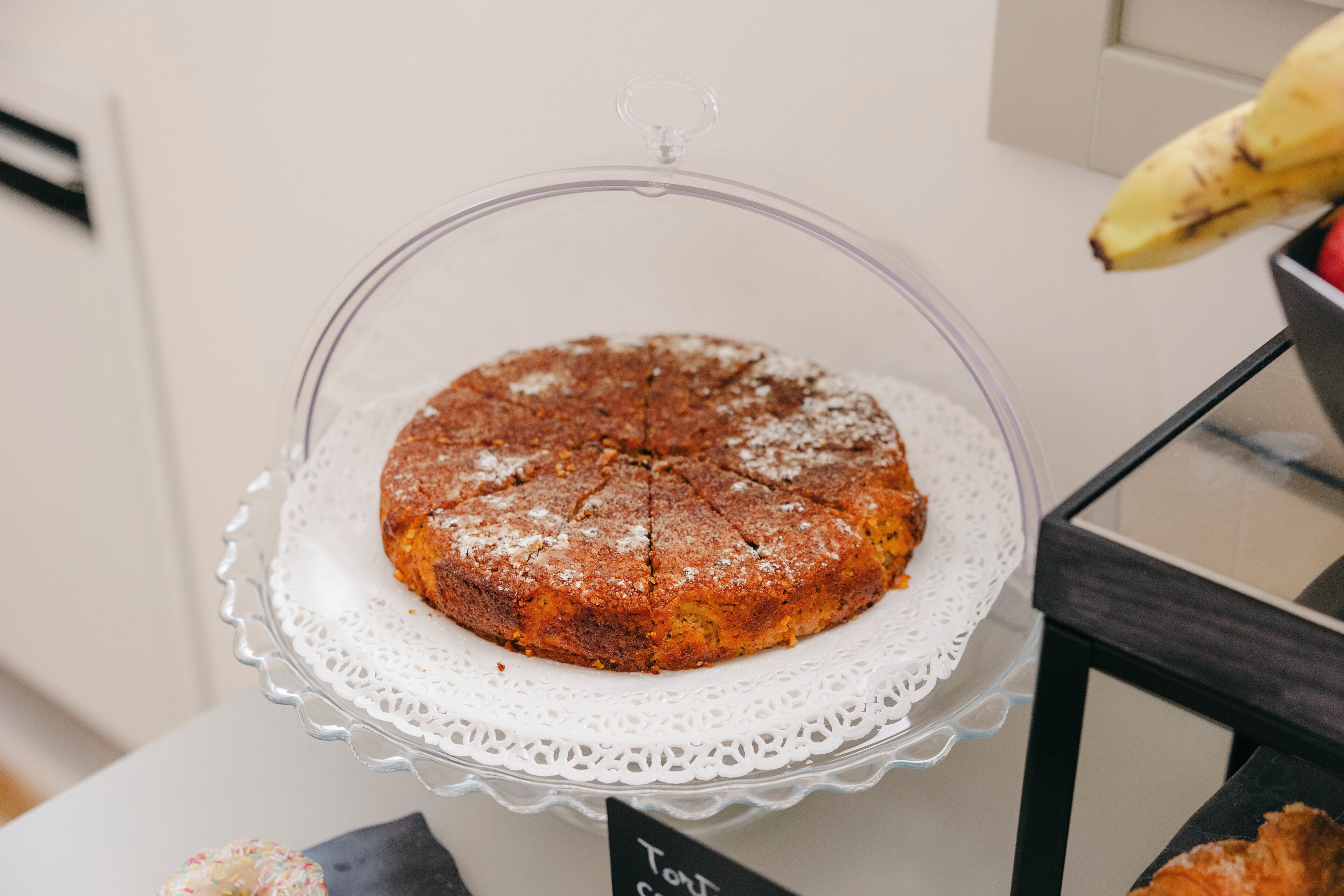 Ein runder Kuchen auf einer Glaskuchengrundplatte mit einer Spitzendecke darunter, auf einem Tisch platziert.