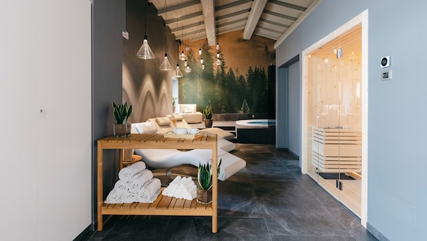 A modern spa area with a wooden table, towels, and plants. There is a seating area with a view of trees and a sauna visible through a glass door. - Villa Turco Berti Luxury Apartments&SPA (Caldiero)