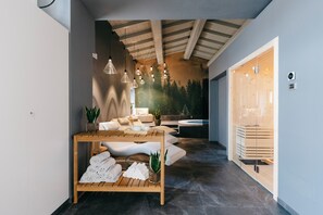 Un moderno espacio de spa con una mesa de madera, toallas y plantas. Hay una zona de estar con vistas a árboles y una sauna visible a través de una puerta de cristal.