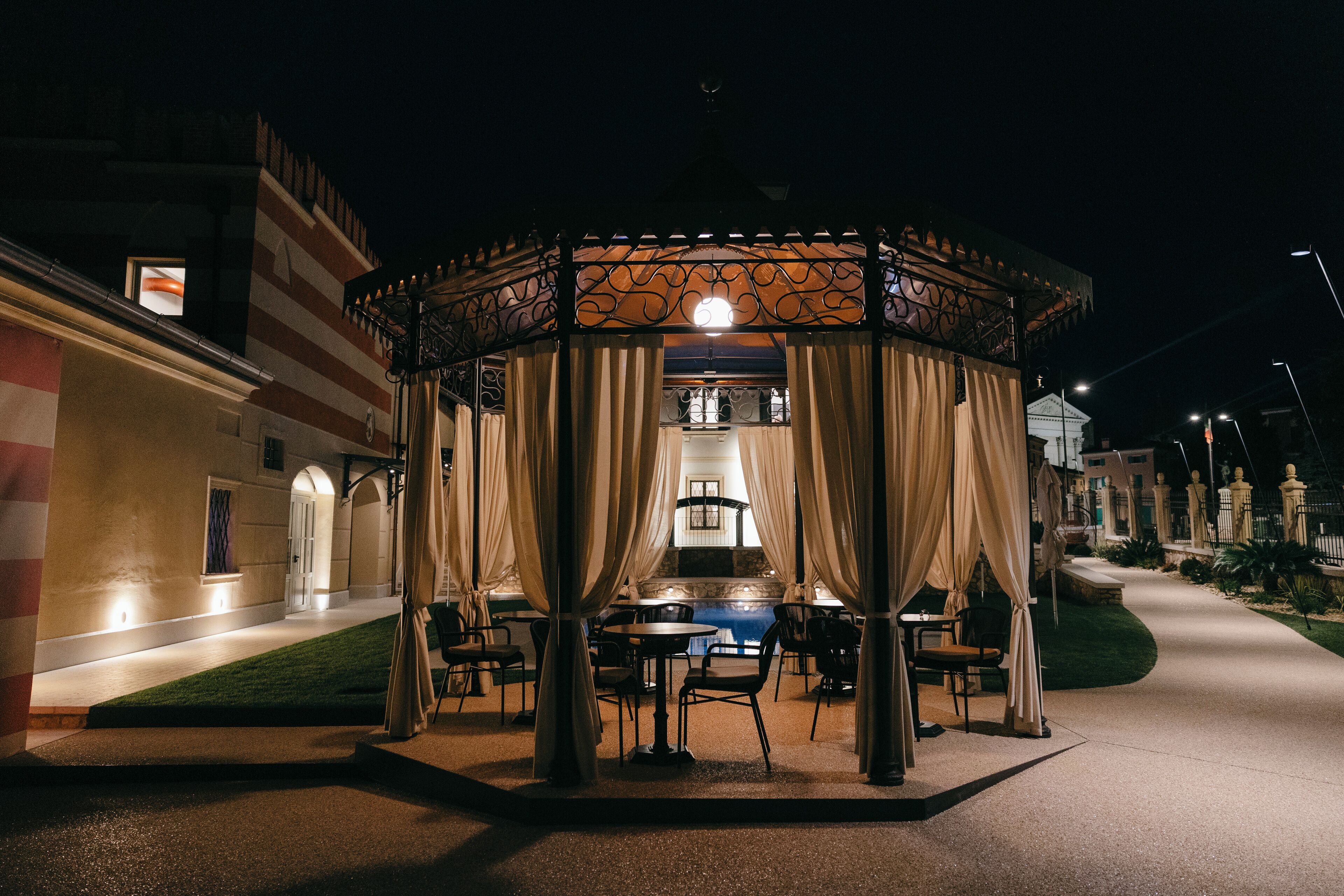 A gazebo with a central light, surrounded by tables and chairs, set against a building facade with a staircase and a garden path.