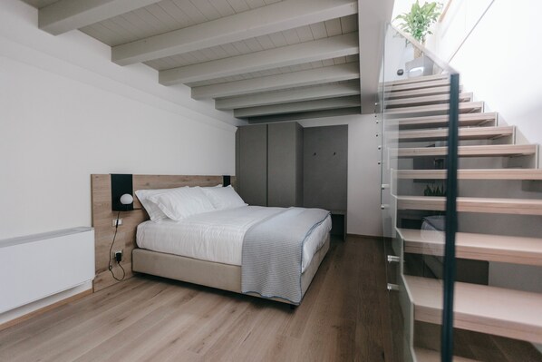 Un dormitorio moderno con piso de madera, cama con ropa blanca, cabecera, mesita de noche con lámpara y una escalera con planta en el descansillo.