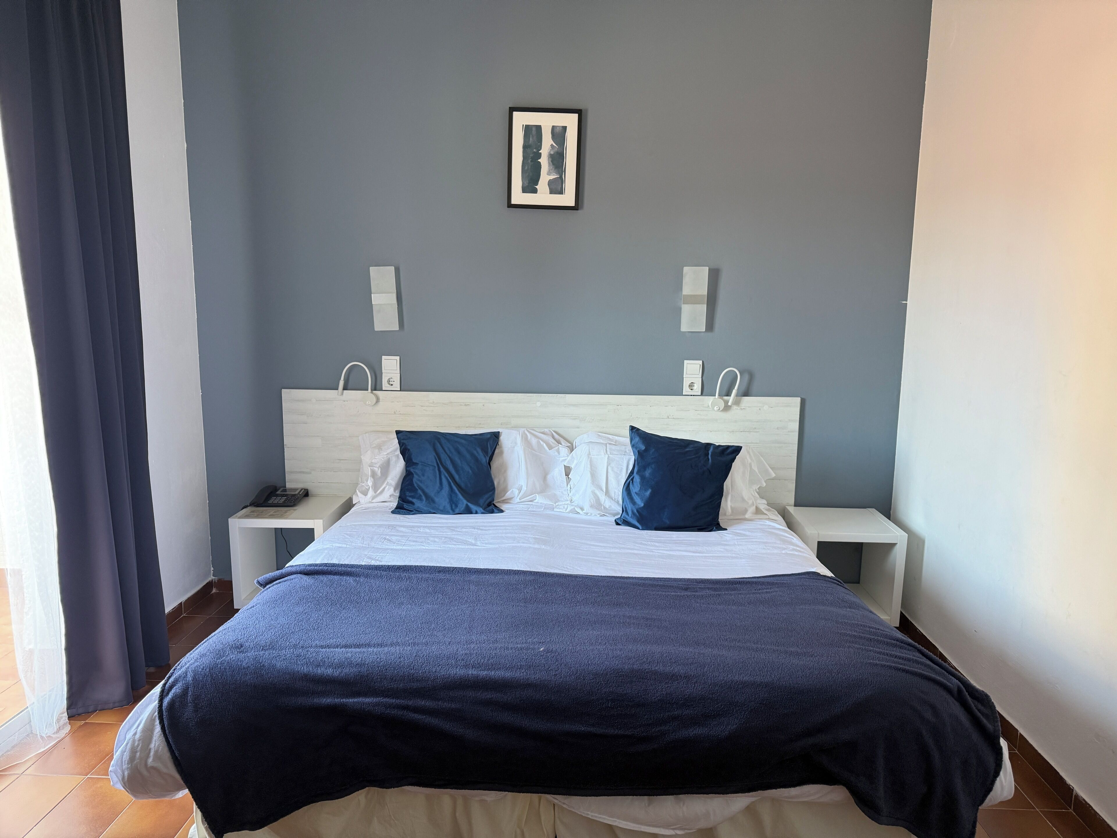 Un lit bien fait, aux couleurs blanches et bleues, avec deux tables de chevet et une tête de lit de couleur claire.