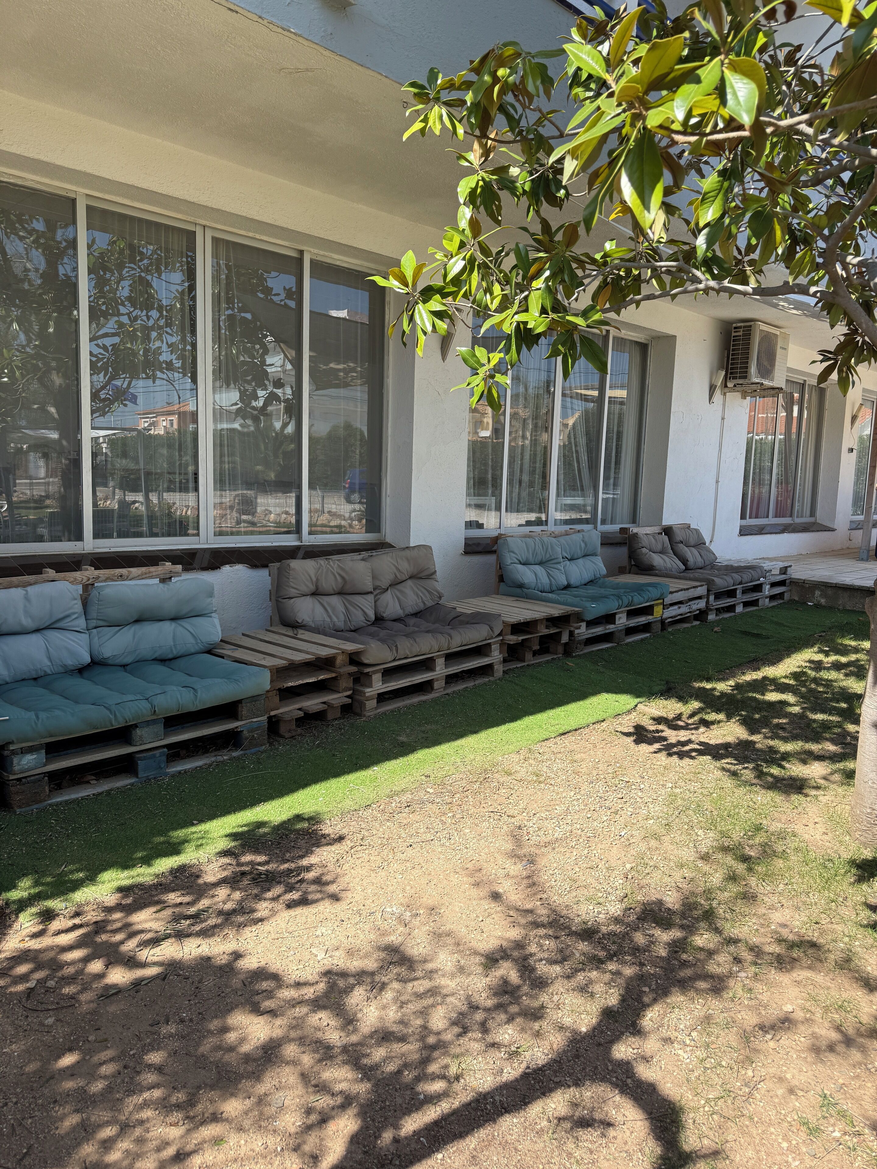 Un patio con zona de estar integrada hecha con palets de madera y cojines, ventanas amplias y un árbol que da sombra.