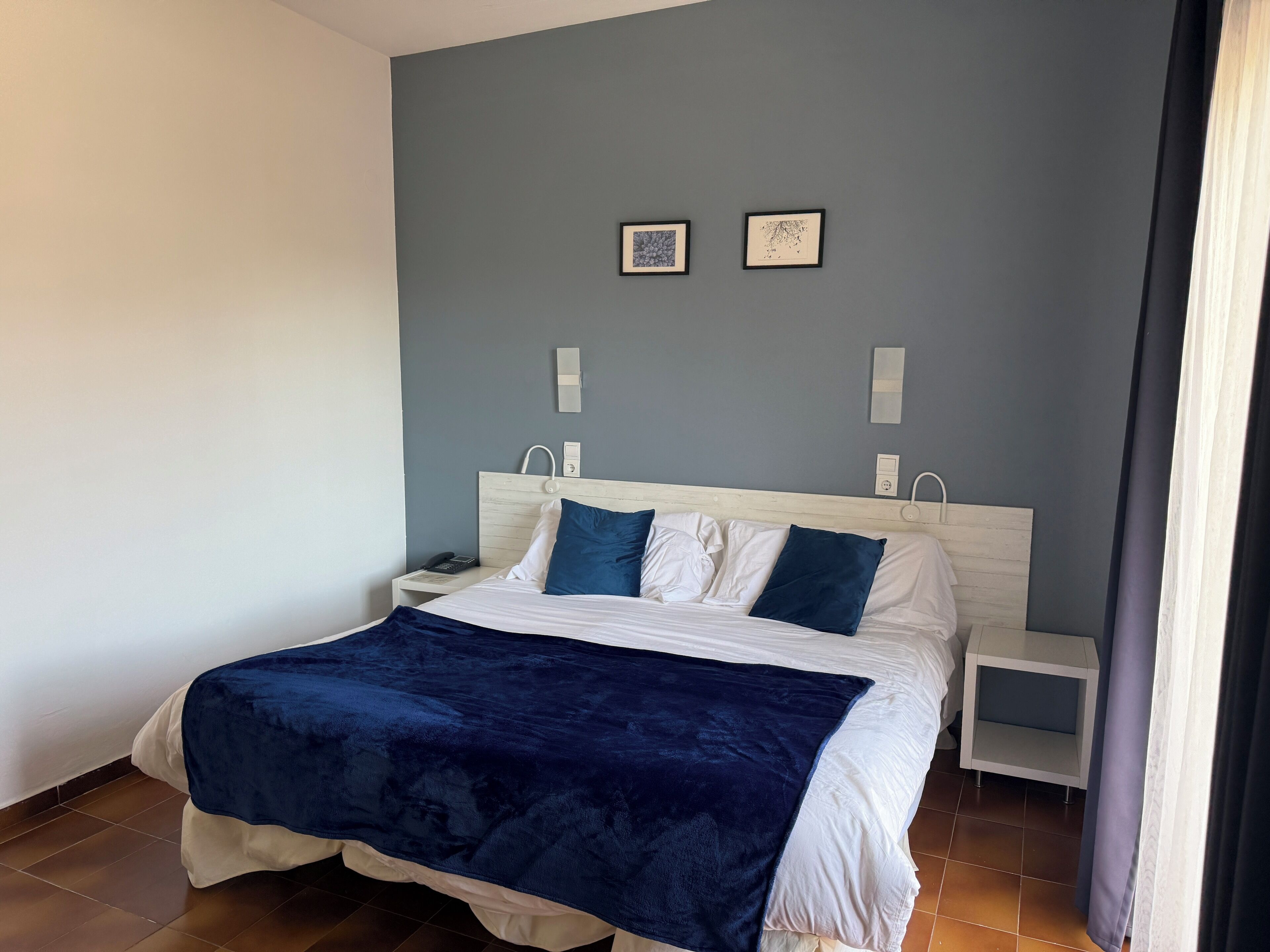 Ein ordentlich bezogenes Bett mit blauer Decke, weißen Kissen und einem Holzkopfstück. Es gibt zwei Nachttische mit Lampen, eine Wandleuchte sowie zwei gerahmte Bilder an der Wand.