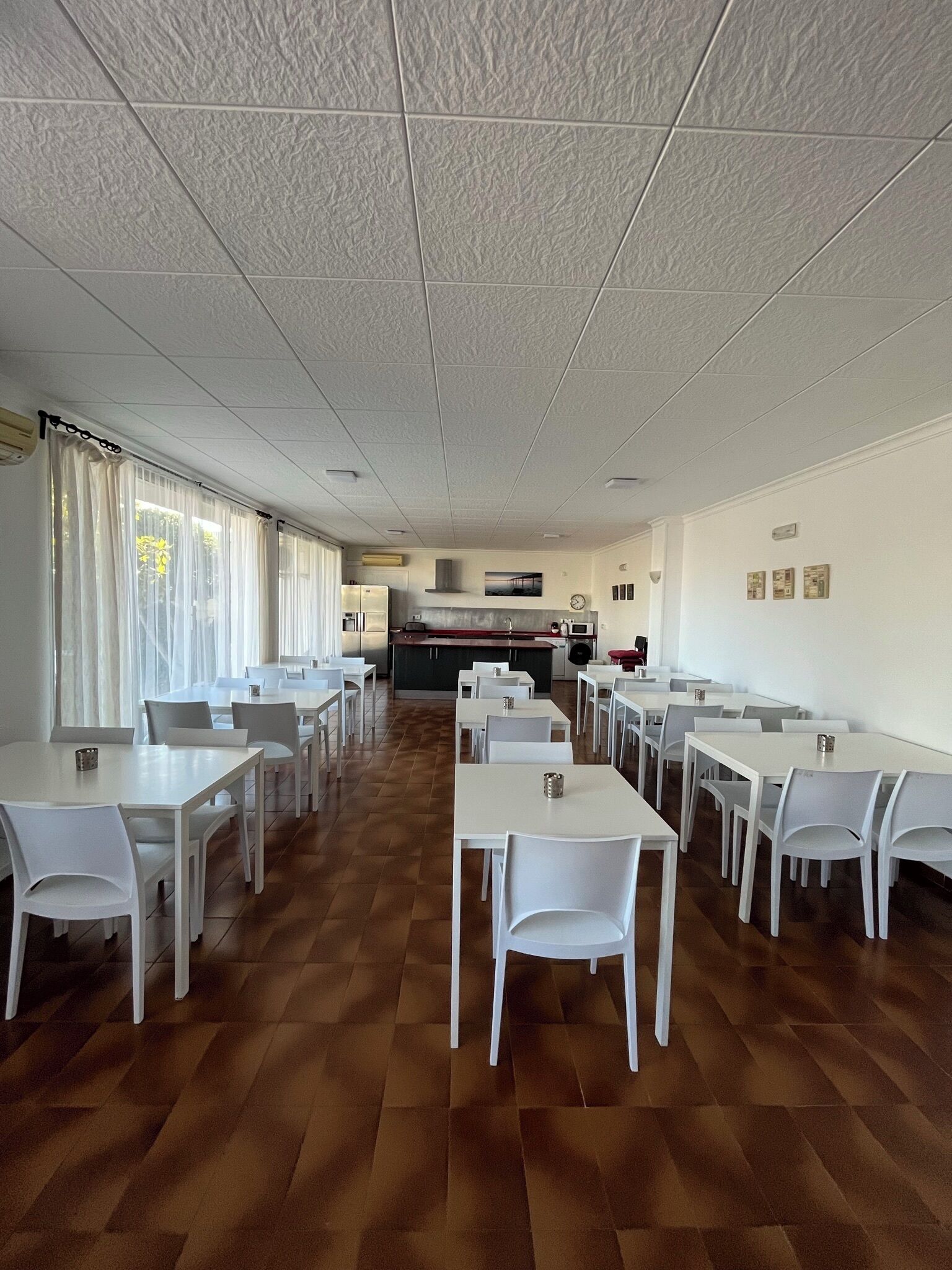Un comedor amplio con mesas y sillas blancas, una cocina al fondo y vistas a través de amplias ventanas.