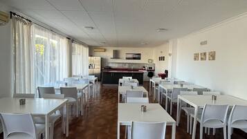 Un comedor amplio con mesas y sillas blancas, una cocina al fondo y vistas a través de amplias ventanas.