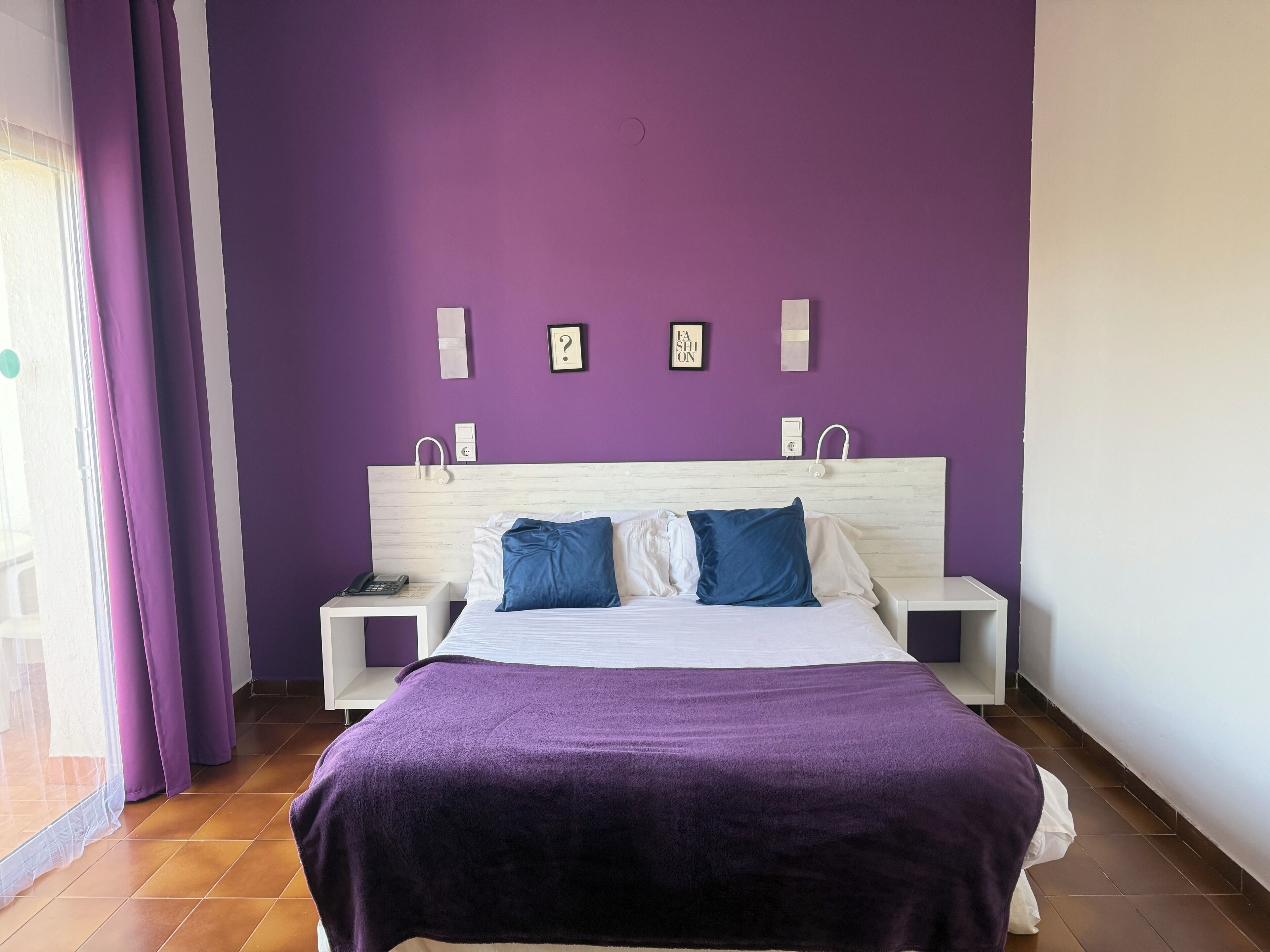 Un dormitorio con una pared morada, una cama con almohadas blancas y azules, y dos mesitas de noche con lámparas.