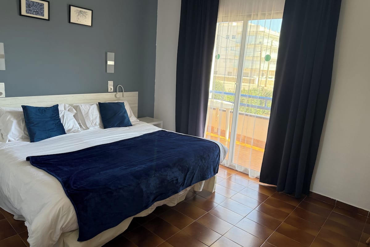 A bedroom with a bed, a nightstand, a wall-mounted light, and a window with curtains.