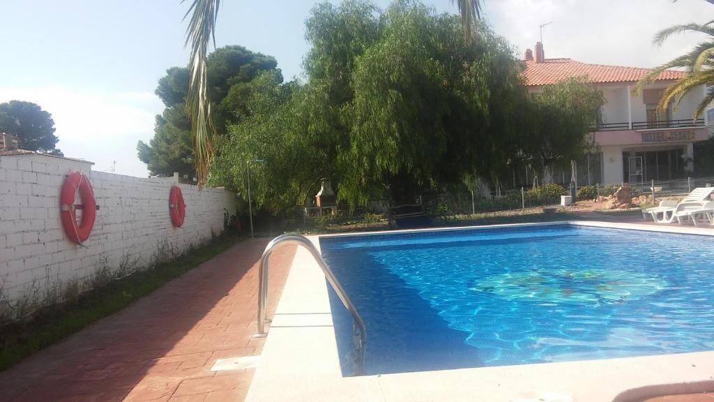 A swimming pool with a white wall and red lifebuoys, surrounded by trees and a building with a red roof.