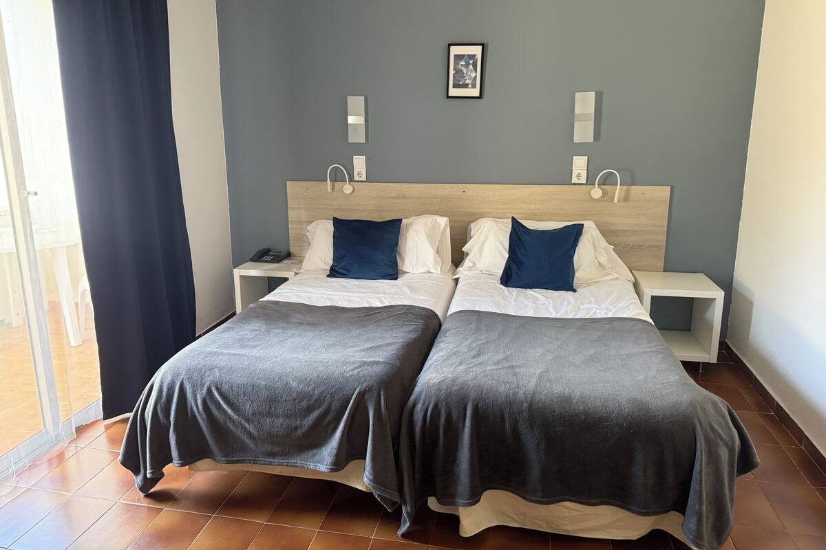 A double bed with a grey bedspread, two white pillows, and a blue accent pillow. There is a bedside table with a phone and a wall-mounted light.