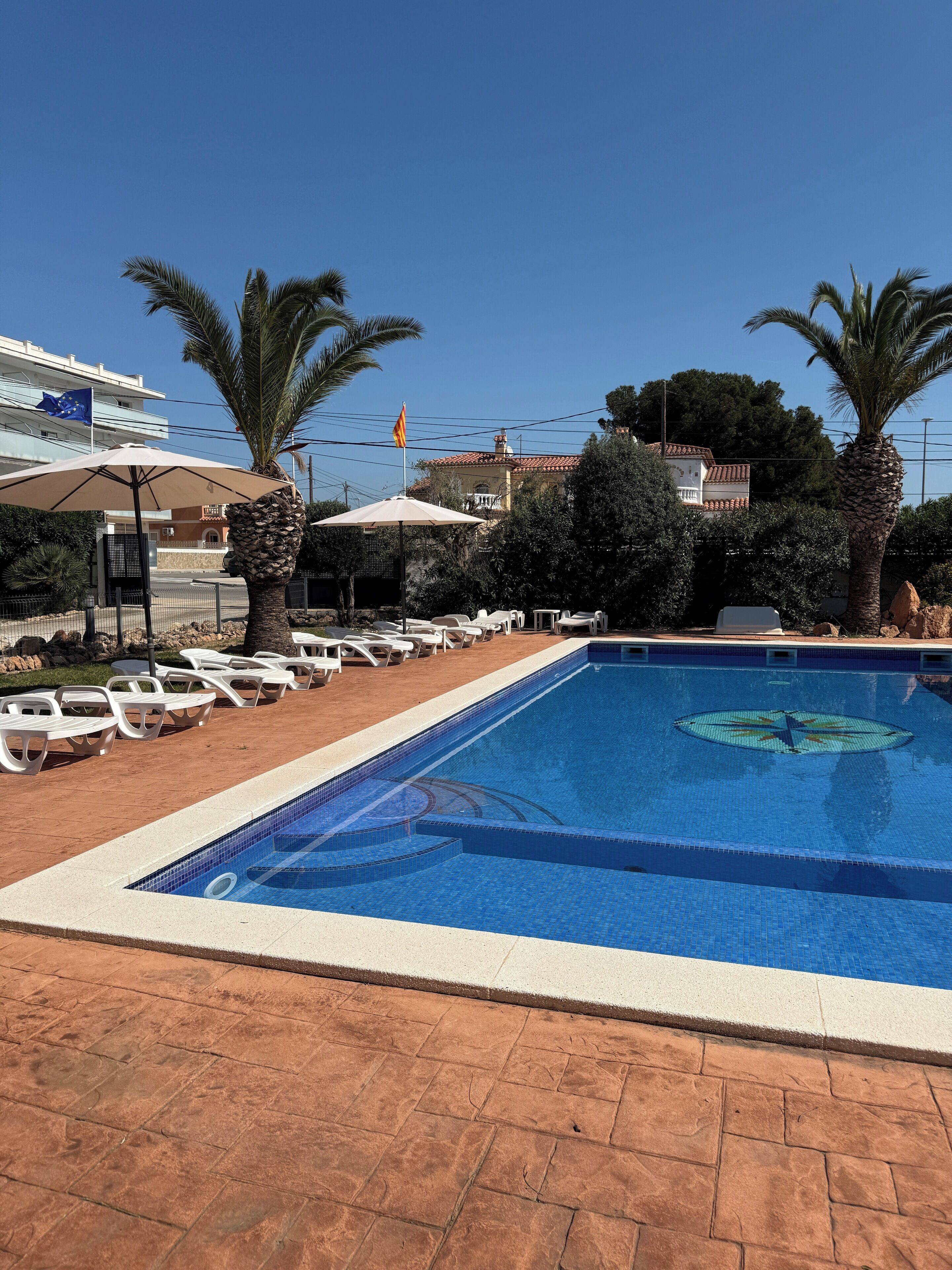 Una piscina con sillas de descanso y sombrillas, rodeada de palmeras y edificios.