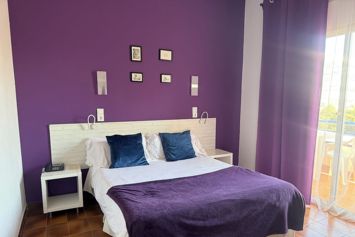 A bedroom with a purple wall, a white bed, and a purple blanket. There are framed pictures on the wall and a nightstand with a phone.