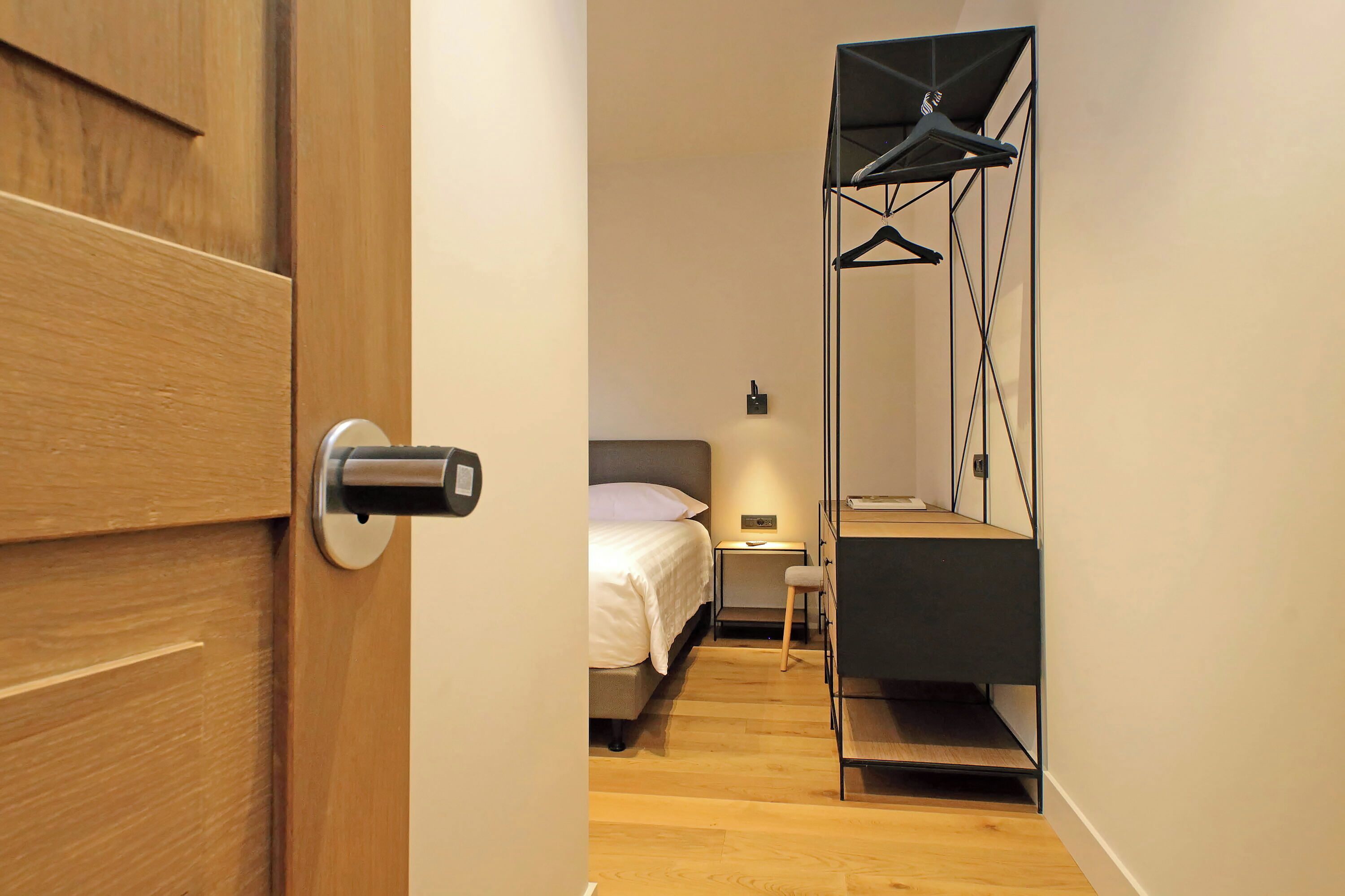 A compact hotel room with a wooden door, a bed with white bedding, a bedside table, a small chair, and a built-in wardrobe with a mirror.