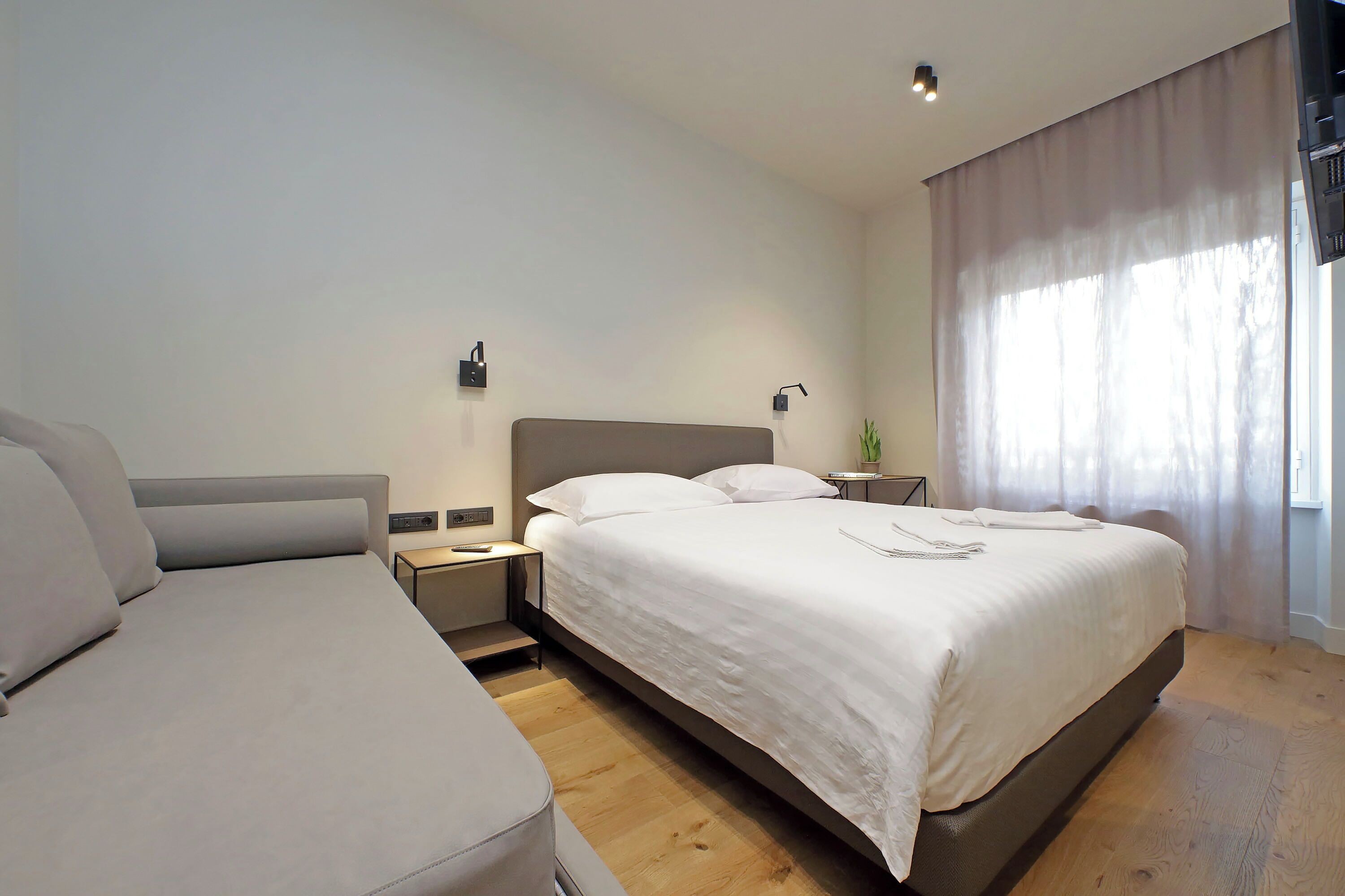 A modern hotel room with a large bed, a smaller sofa, a bedside table, and a nightstand.