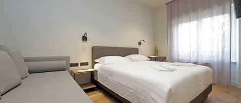 A modern hotel room with a large bed, a smaller sofa, a bedside table, and a nightstand.