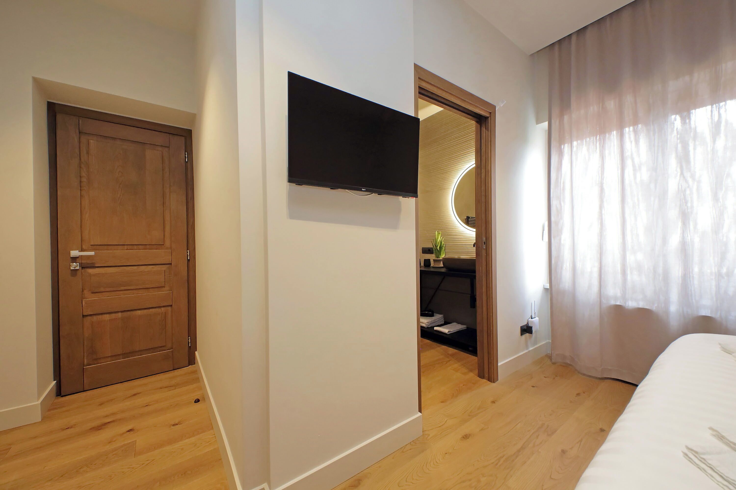 A hotel room with a wooden door, a mounted TV, a bed with white bedding, and a large window with sheer curtains.