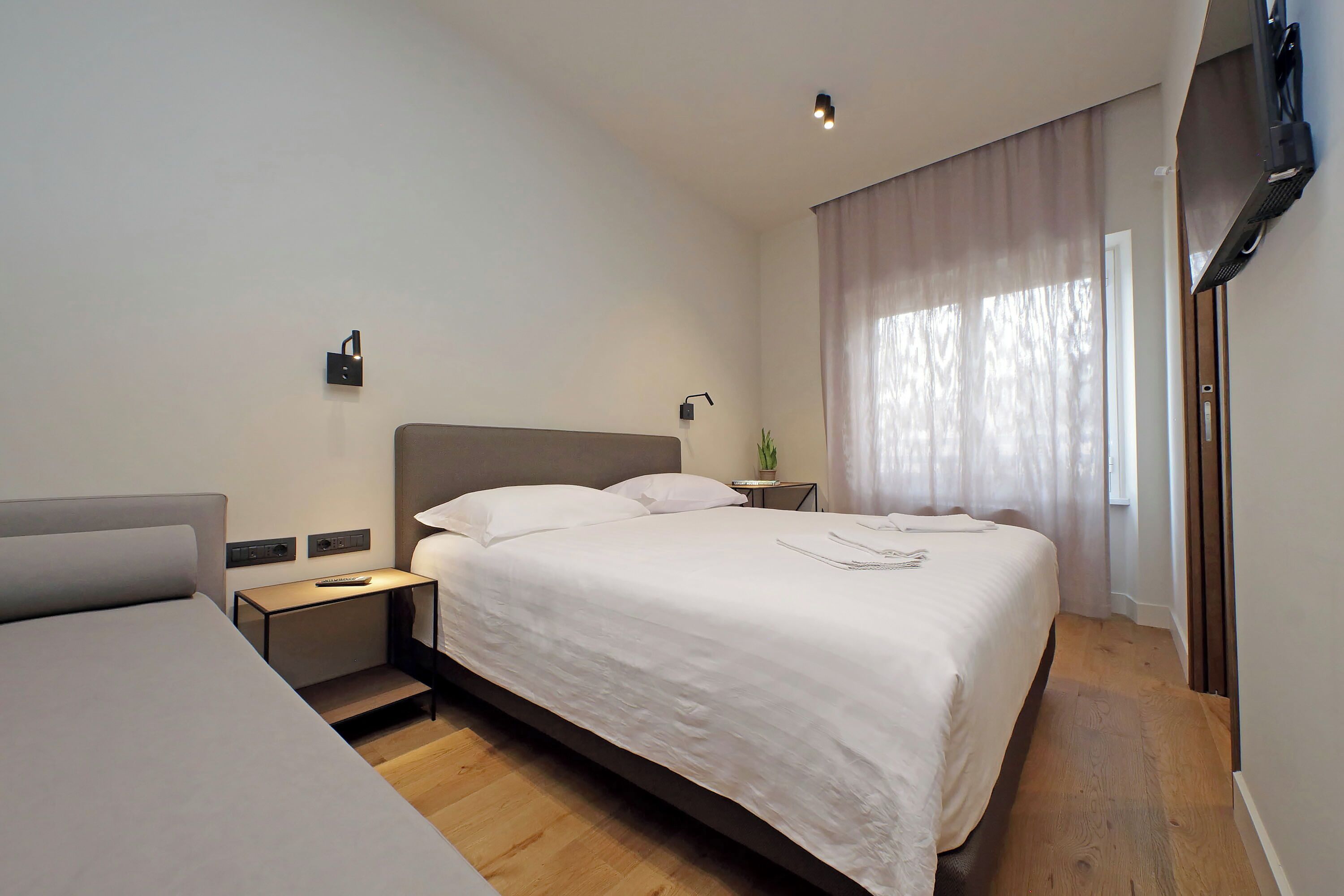 A modern hotel room with a large bed, bedside tables, a small table, and a TV mounted on the wall.