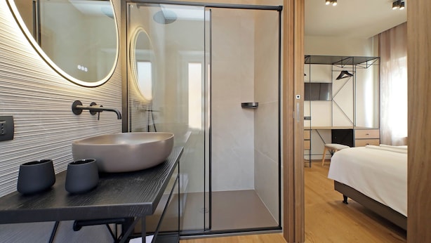 A modern bathroom with a glass shower enclosure, a vanity with a large sink, and a bed in the adjacent room. - The Place Gallia (Rome)