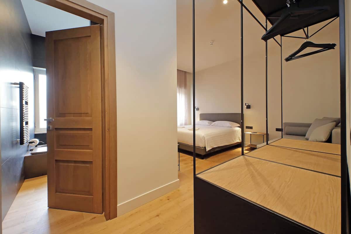 A modern bedroom with a wooden door, a bed with a grey headboard, and a large mirror reflecting the room.