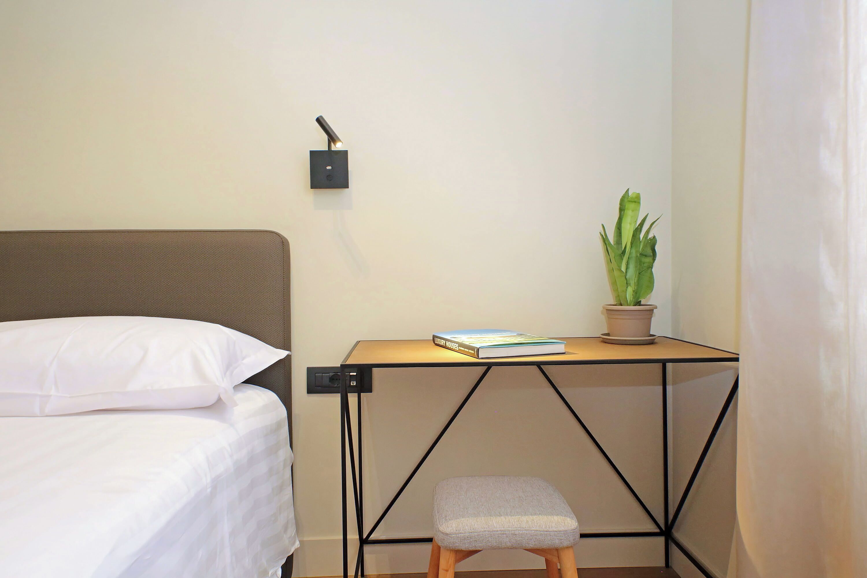 A neatly made bed with white linens, a small wooden stool, a table with a book and a potted plant, and a wall-mounted light fixture.