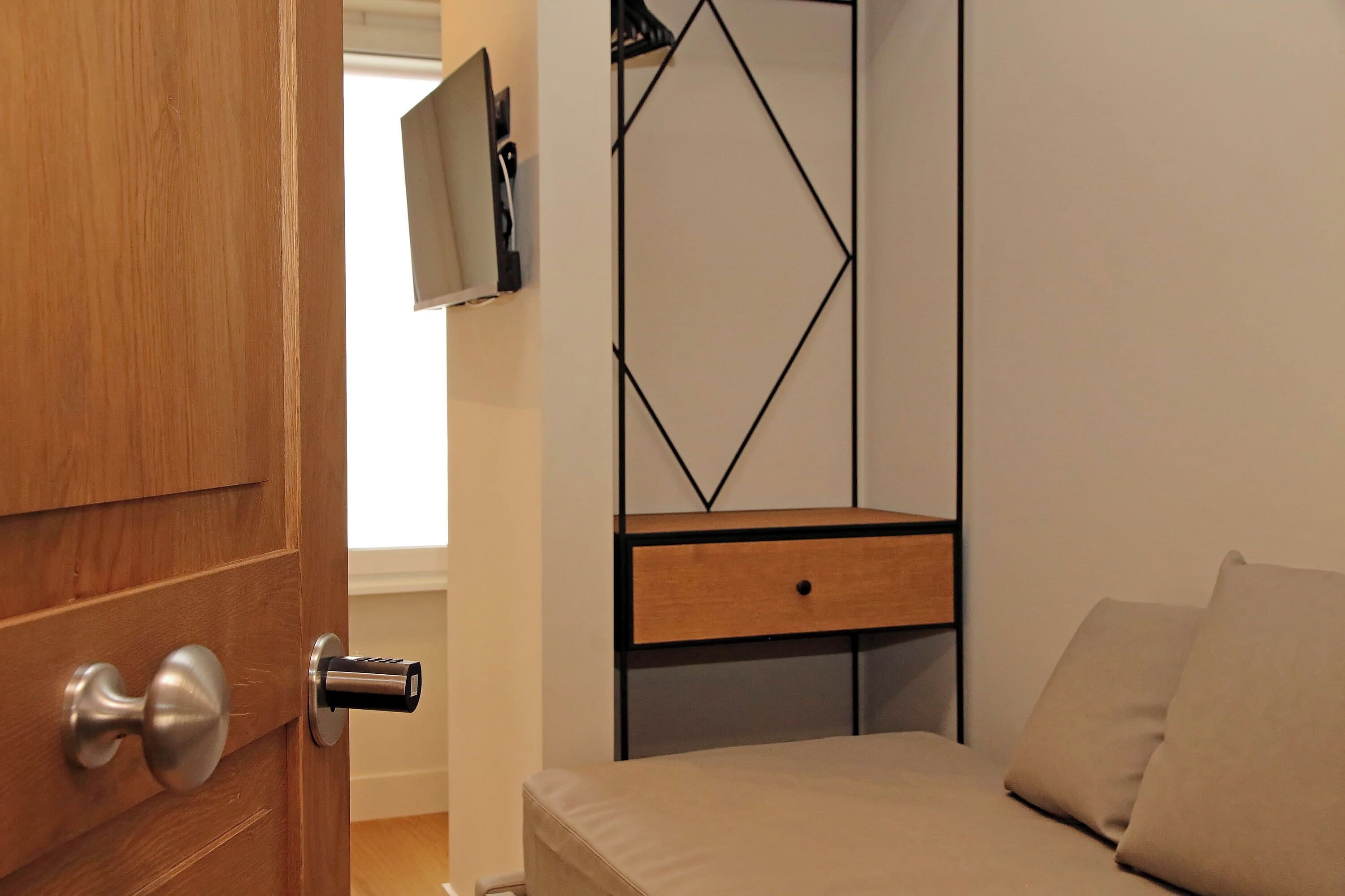 Un dormitorio con puerta de madera, una cama con ropa de cama color crema, una mesita de noche de madera y un televisor montado en la pared.