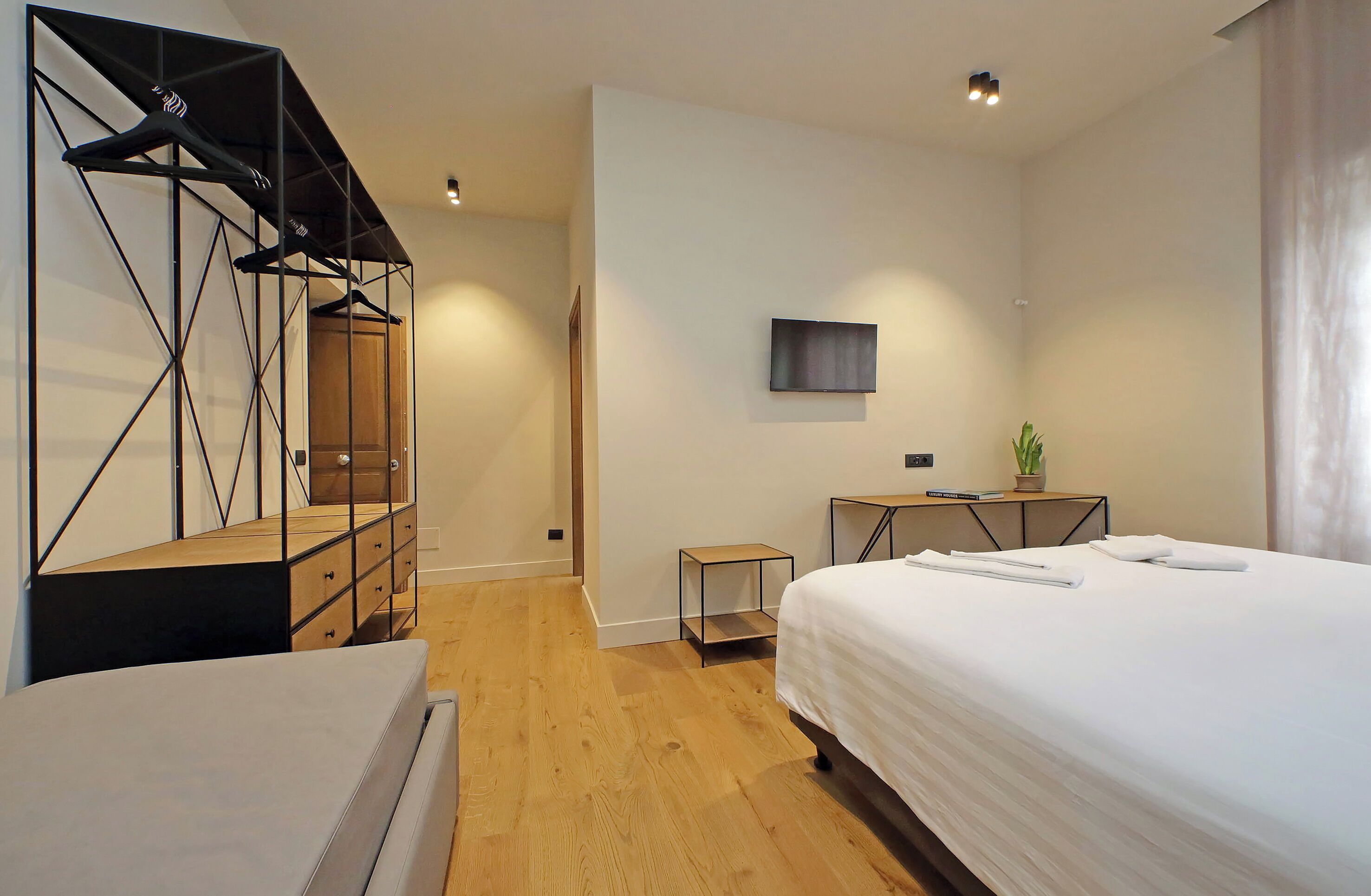 A modern hotel room with a wooden floor, a bed with white linens, a TV mounted on the wall, a small table with a plant, and a unique geometric shelving unit.
